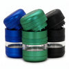 Three metal herb grinders in blue, green, and black, arranged together on a white background. Each grinder has a clear glass or plastic chamber in the middle.