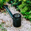 A dark green Jane West Twenties Collection Glas Hand Pipe tanding upright on a gravel path surrounded by lush green plants.