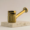 A polished gold-colored metal pipe with a textured, diamond-patterned body and a curved stem, displayed on a light gray marble block against a neutral light gray background.
