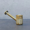 A polished brass smoking pipe with a textured diamond-patterned bowl and a straight, angled stem, resting on a dark gray surface against a gray concrete wall.