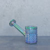 An iridescent glass smoking pipe with a diamond-textured bowl and stem, showing a holographic rainbow sheen, set against a neutral gray background.