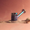 An iridescent metallic smoking pipe or torch lighter with a textured cylindrical body and angled stem, resting on sand with seashells and mist against a warm pink background.
