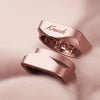 Two rose gold rings with a modern geometric design, one featuring the engraved script name 'Krush' and the other displaying a 3D geometric cutout interior, resting on a soft, textured pink surface.