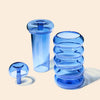 Three translucent blue glass smoking accessories on a warm beige background: a small dome-shaped piece, a tall cylindrical piece, and a stacked set of ribbed circular pieces.