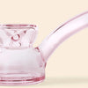 A close-up of a transparent pink glass water pipe by NWTN Home with a curved mouthpiece, shown against a solid pale beige background.